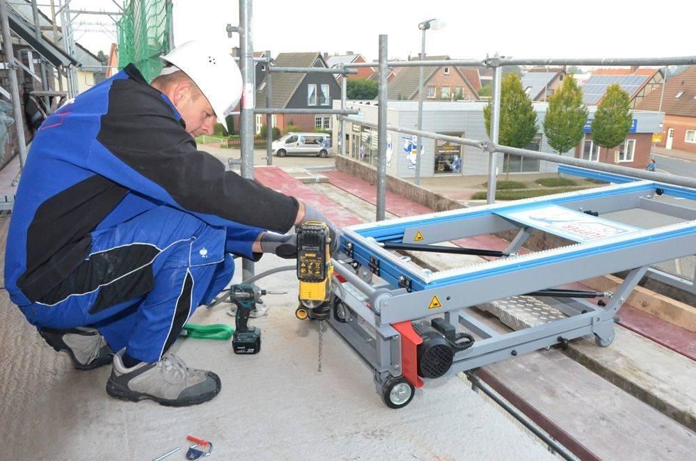 Mann bohrt auf Gerüst; verwendet tragbaren Arbeitstisch. Blauer Overall, weißer Schutzhelm, städtische Umgebung.
