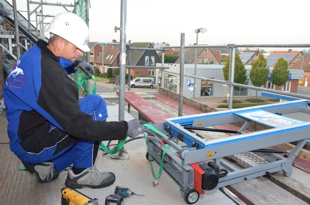 Bauarbeiter auf einem Gerüst sichert einen blauen Karren mit Gurten. Er trägt einen Schutzhelm.