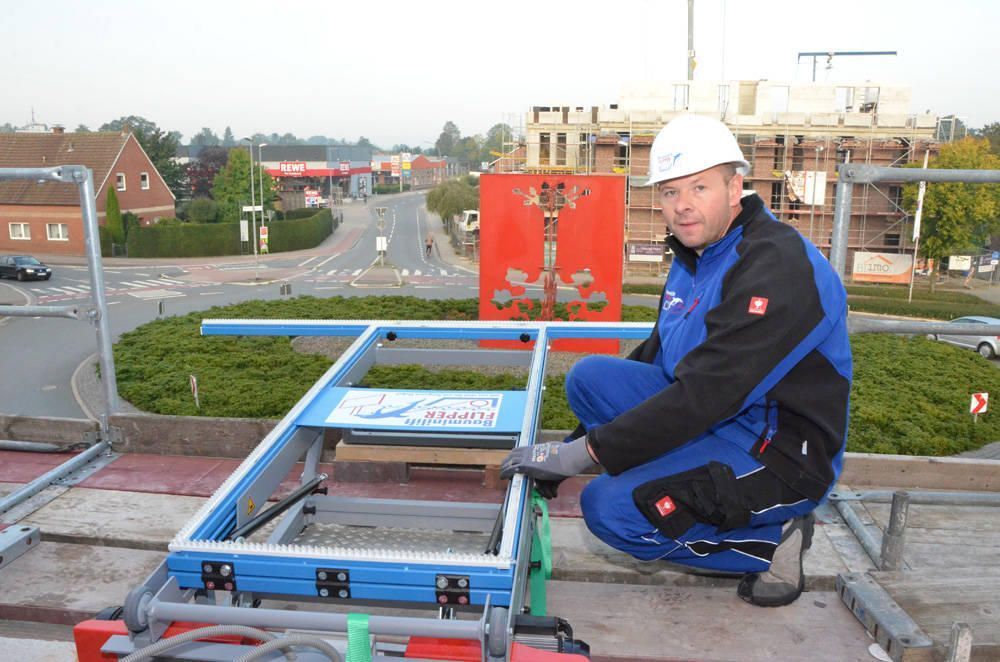 Bauarbeiter kniet neben einem blau-silbernen Gerüst und trägt einen weißen Helm auf einem Dach mit Blick auf eine Stadt.