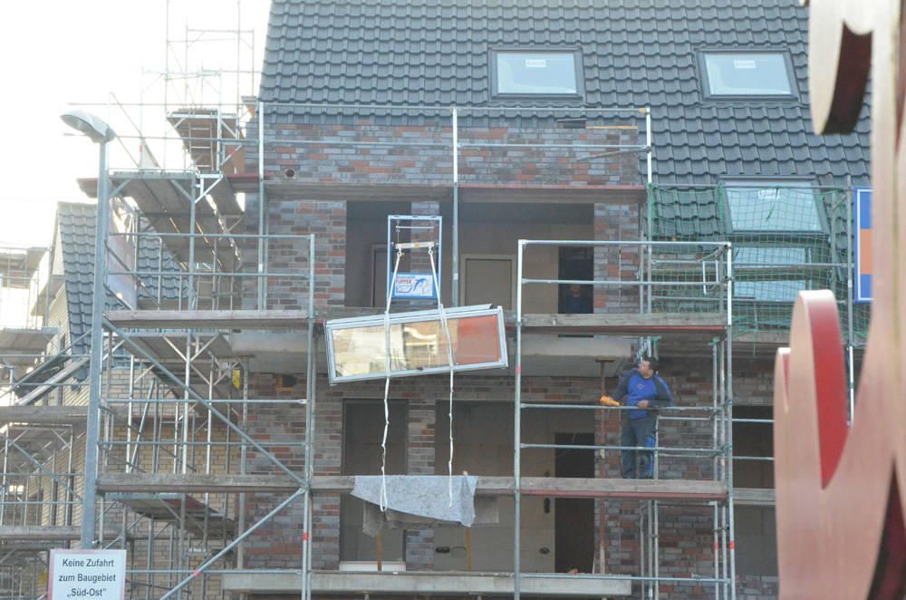 Bauarbeiter auf Gerüst, hebt Fensterrahmen an einem Gebäude an, Ziegelfassade, graues Dach.