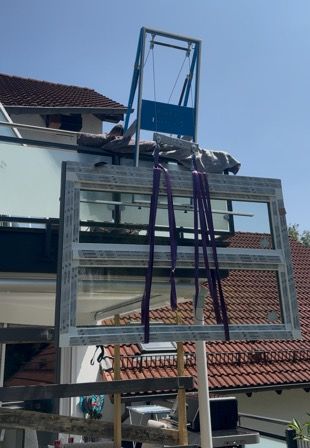 Ein großer Fensterrahmen wird an einem sonnigen Tag von einem Kran angehoben. Der Kran ist blau und silber.