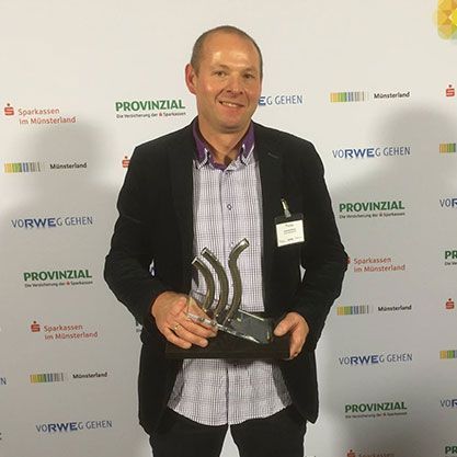 Mann im Anzug hält eine Auszeichnung in der Hand, lächelt und steht vor einem Banner mit Logos.