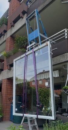 Ein großes Fenster wird mit einem Kran auf einen Balkon gehoben. Blauer Kran, violette Gurte und weißer Fensterrahmen.