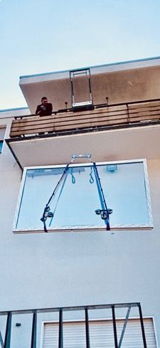 Ein großes Fenster wird mit Gurten in ein Gebäude gehoben, eine Person steht auf dem Balkon darüber.