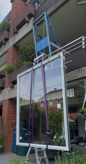 Ein großes Fenster wird mit einem Hebezeug auf einen Balkon gehoben. Lilafarbene Gurte sichern es an einem Ständer.