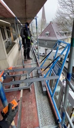 Ein Bauarbeiter auf einem Gerüst mit einer Fernbedienung in der Hand. Im Hintergrund sind Gebäude und ein Kirchturm zu sehen.
