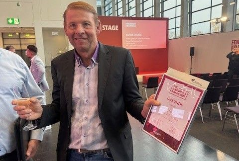 Ein Mann im Anzug an einem Konferenzstand, lächelnd und mit einem Tablet in der Hand, hinter ihm ein rotes Schild.