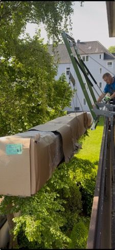 Ein Mann räumt mit einem Kran ein großes, verpacktes Paket von einem Balkon. Im Hintergrund sind grüne Bäume und ein Gebäude.