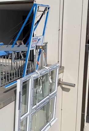 Blauer Kran hebt ein Fenster mit weißem Rahmen vor einem Gebäude an; Fenster wird installiert/entfernt.