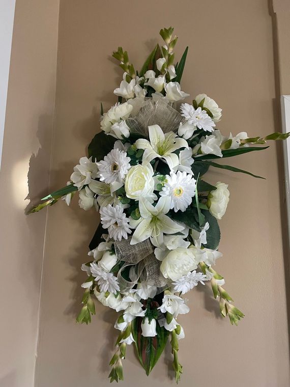 Composition florale funéraire blanche et verte avec des lys et des chrysanthèmes sur un mur beige