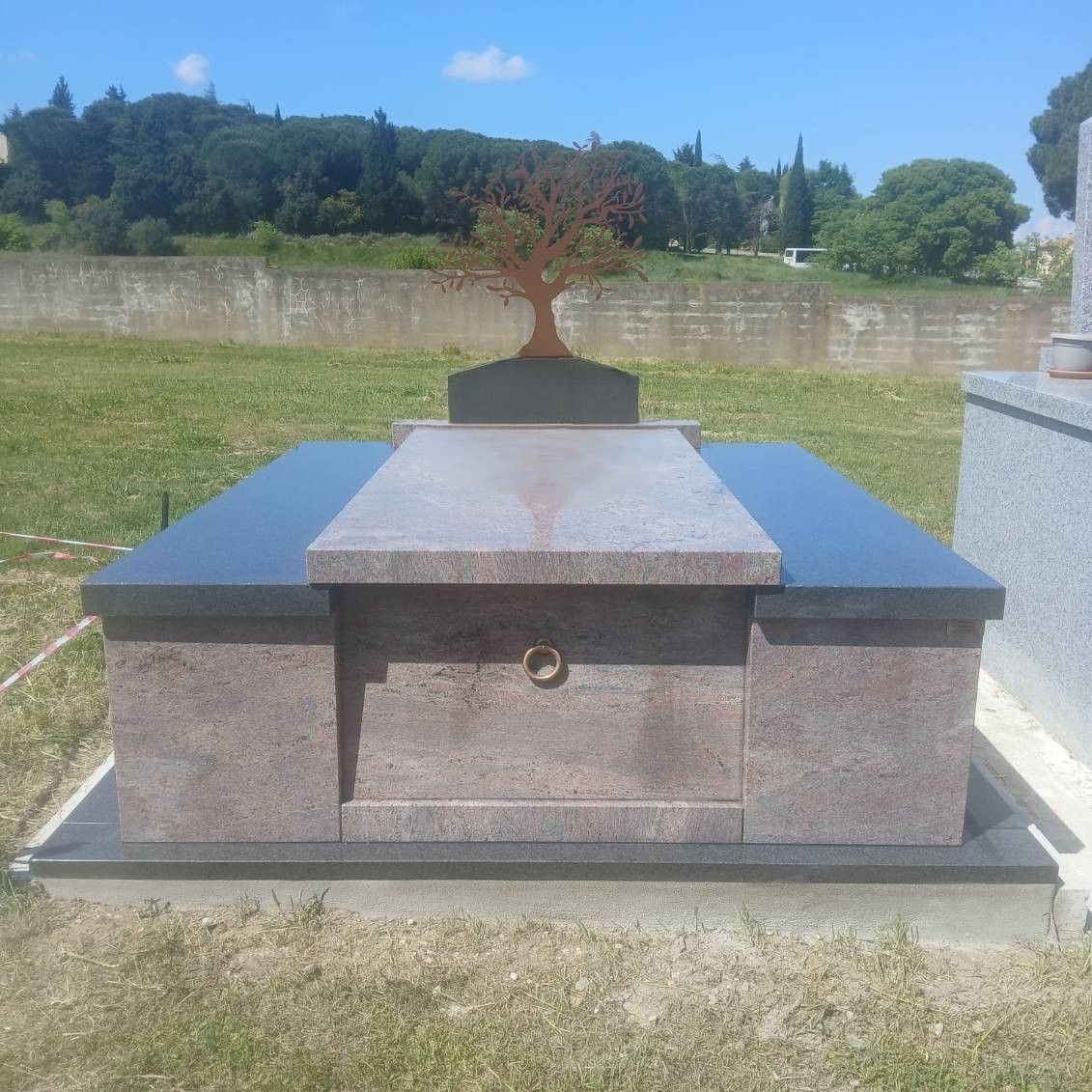 Tombeau commémoratif en pierre avec un petit bonsaï, situé dans un cimetière verdoyant sous un ciel bleu limpide.