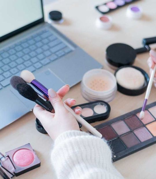 Una mujer sostiene pinceles de maquillaje frente a una computadora portátil.