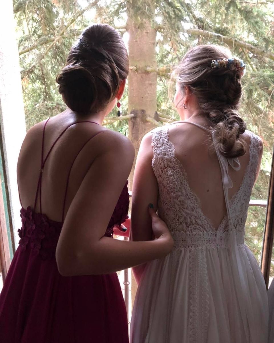 Dos mujeres con vestidos formales mirando por una ventana. Una de color burdeos, la otra de encaje blanco.