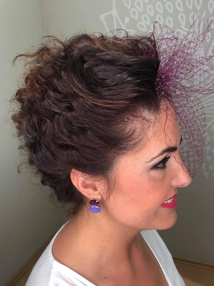 Mujer con cabello castaño y rizado, velo morado, aretes morados y camisa blanca.