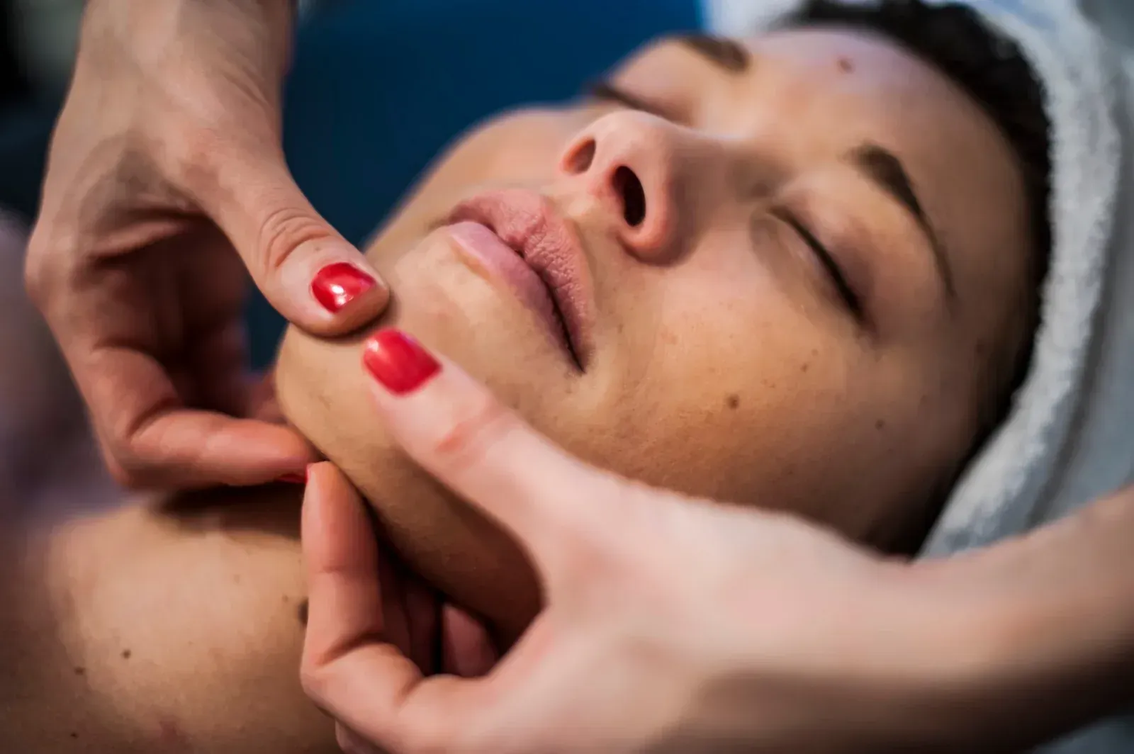 Manos masajeando la barbilla de una persona durante un tratamiento facial. La persona tiene los ojos cerrados; esmalte de uñas rojo.