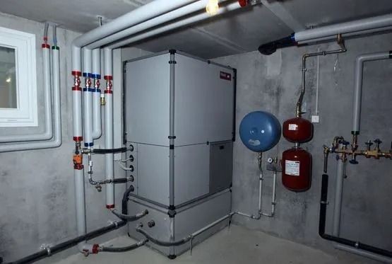 Un système de chauffage dans un sous-sol en béton gris. Il comprend une grande unité centrale, des tuyaux et des réservoirs.