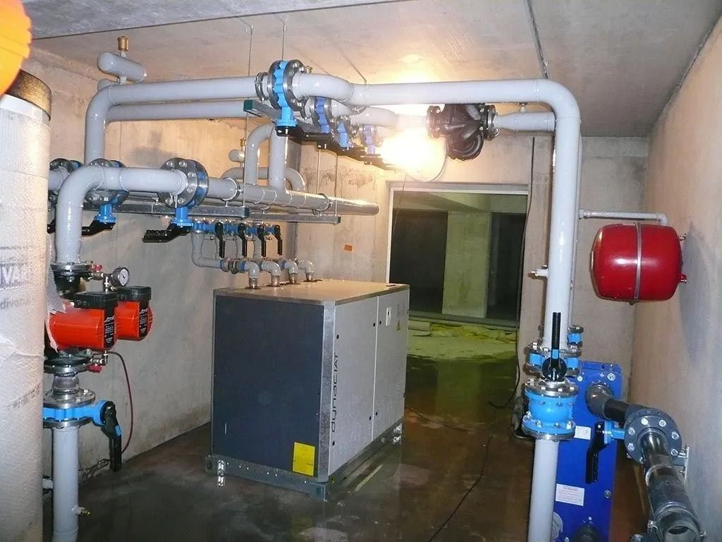 Tuyaux, vannes, pompes et un grand boîtier gris dans un local technique. La scène se déroule à l'intérieur, avec un réservoir rouge et une lumière vive.