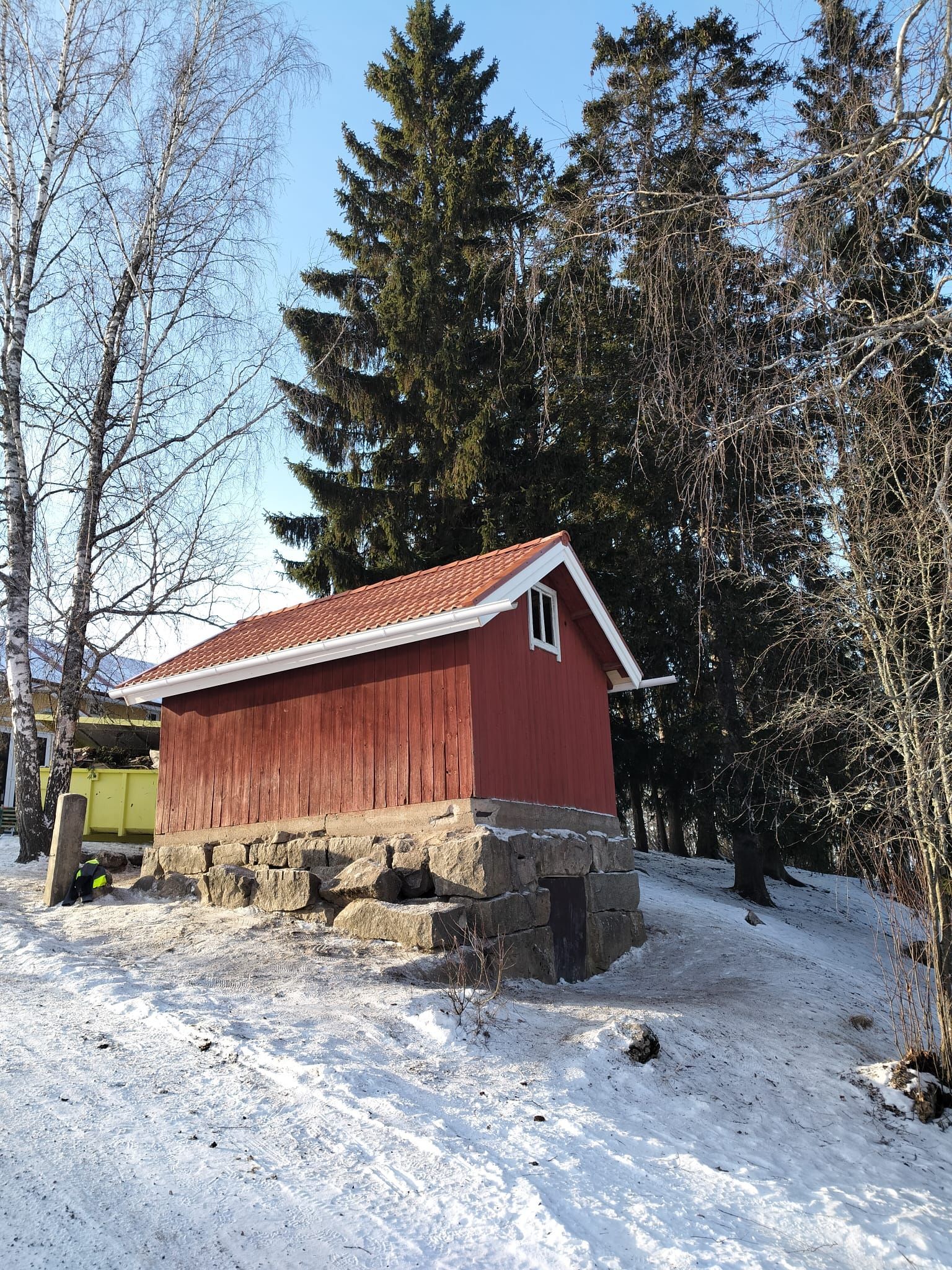 Pieni punainen puinen mökki, jossa on valkoreunainen katto, seisoo kivijalustoilla lumisessa, metsäisessä maisemassa.