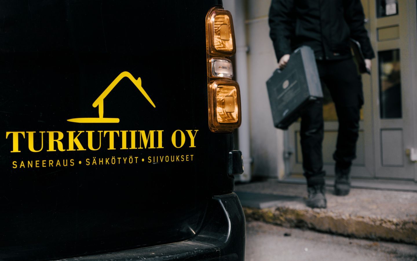 Musta pakettiauto, jossa on keltainen βTURKUTIMI OYβ -logo, salkkua kantavan henkilΓΆn vieressΓ€ ovensuussa