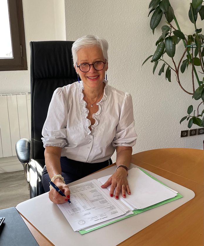 Femme aux cheveux gris courts, portant des lunettes, souriante en signant un document à son bureau. Cadre de bureau.