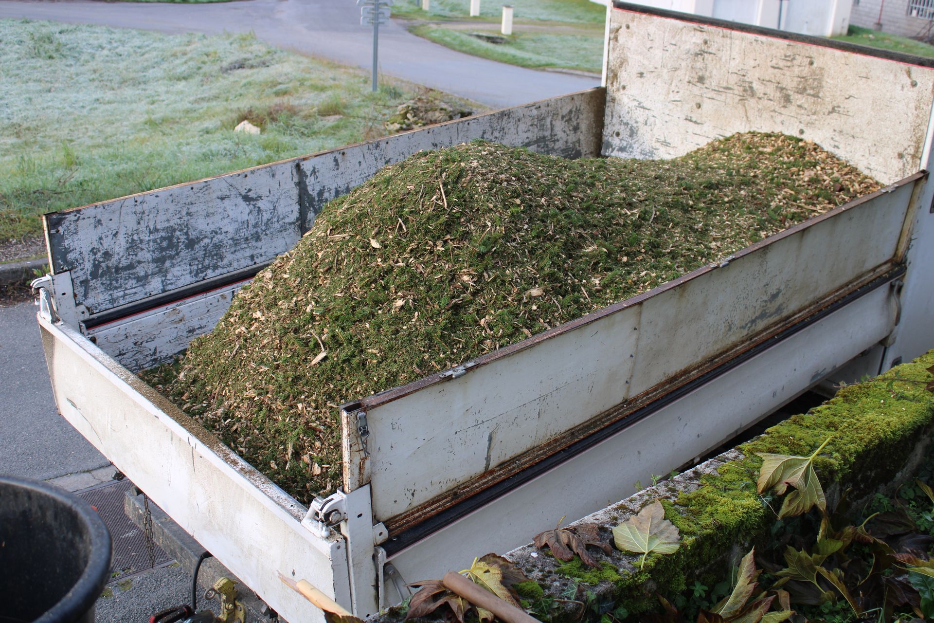 Grande remorque métallique remplie de déchets verts de jardin, garée à côté d'une allée.