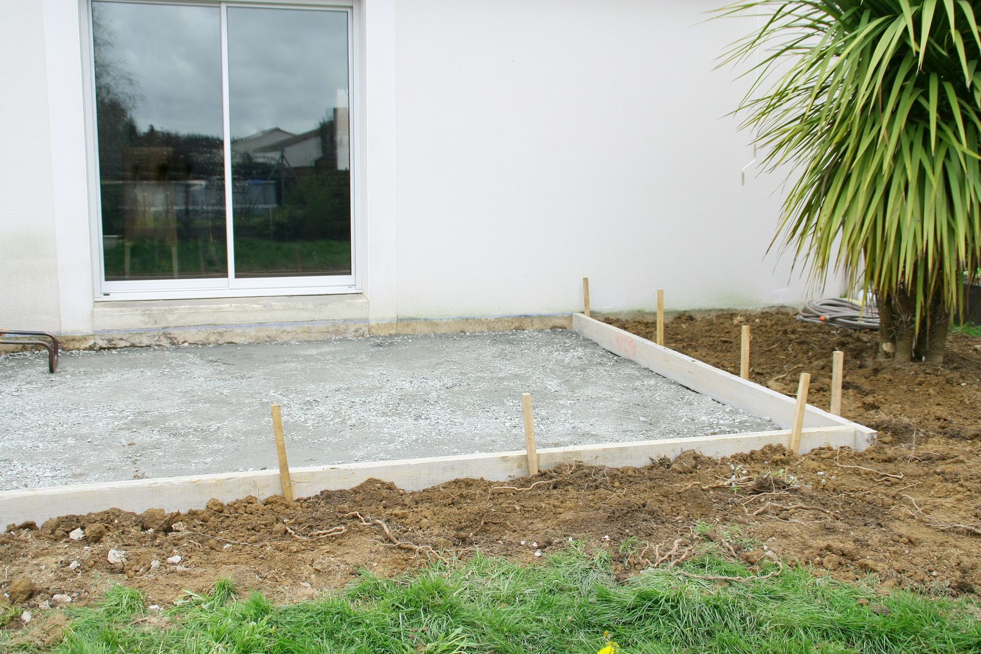 Dalle de terrasse en béton en construction à côté d'une maison, entourée de gravier, de piquets en bois et de terre.