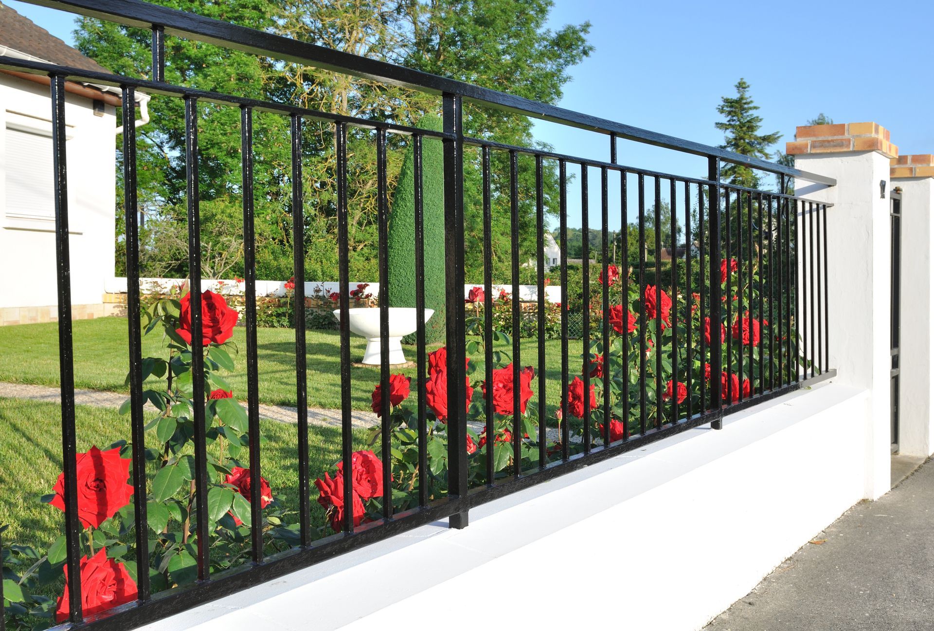 Clôture en métal noir bordant un jardin paysager avec des fleurs rouges sous un ciel bleu.