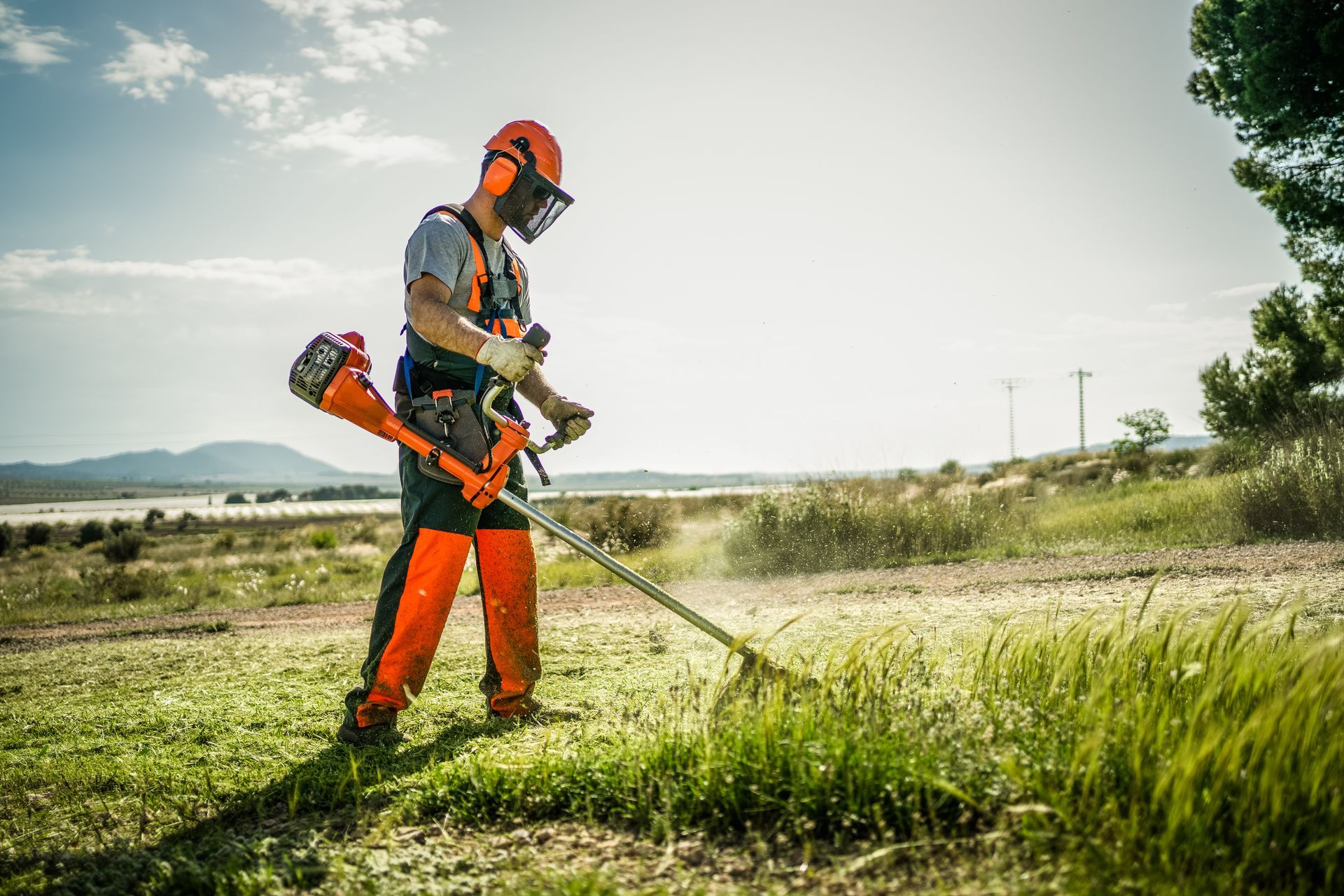 Un ouvrier en tenue de sécurité orange utilise un coupe-bordures dans un champ herbeux sous un ciel éclatant.