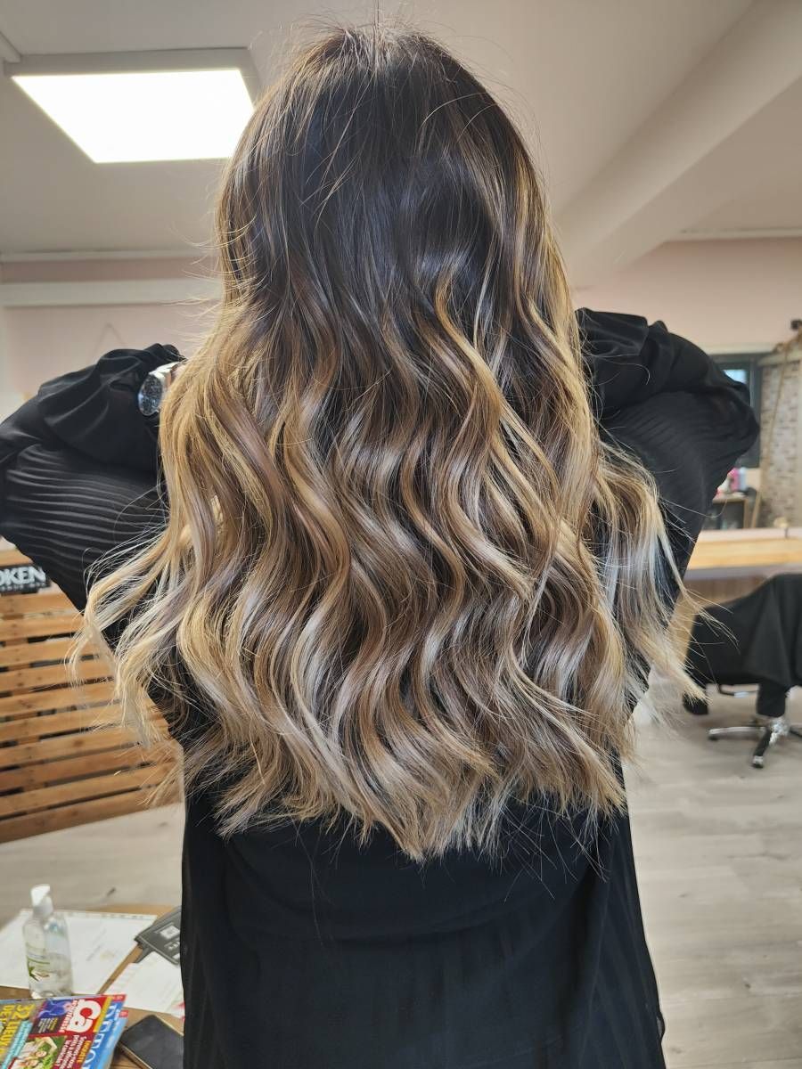 Longs cheveux ondulés ombrés, du brun au blond, vus de dos sur un fauteuil de salon de coiffure