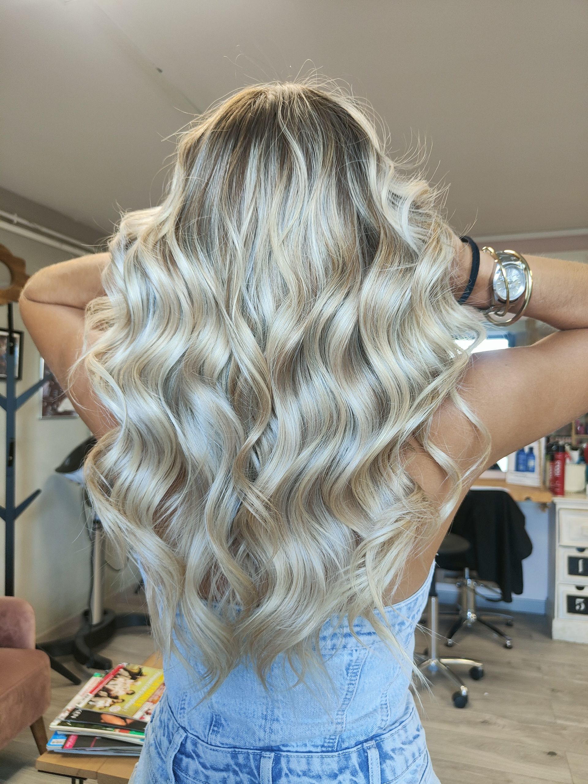 Femme aux longs cheveux blonds ondulés, vue de dos, vêtue d’une robe bleue, debout à l’intérieur, les mains dans les cheveux.