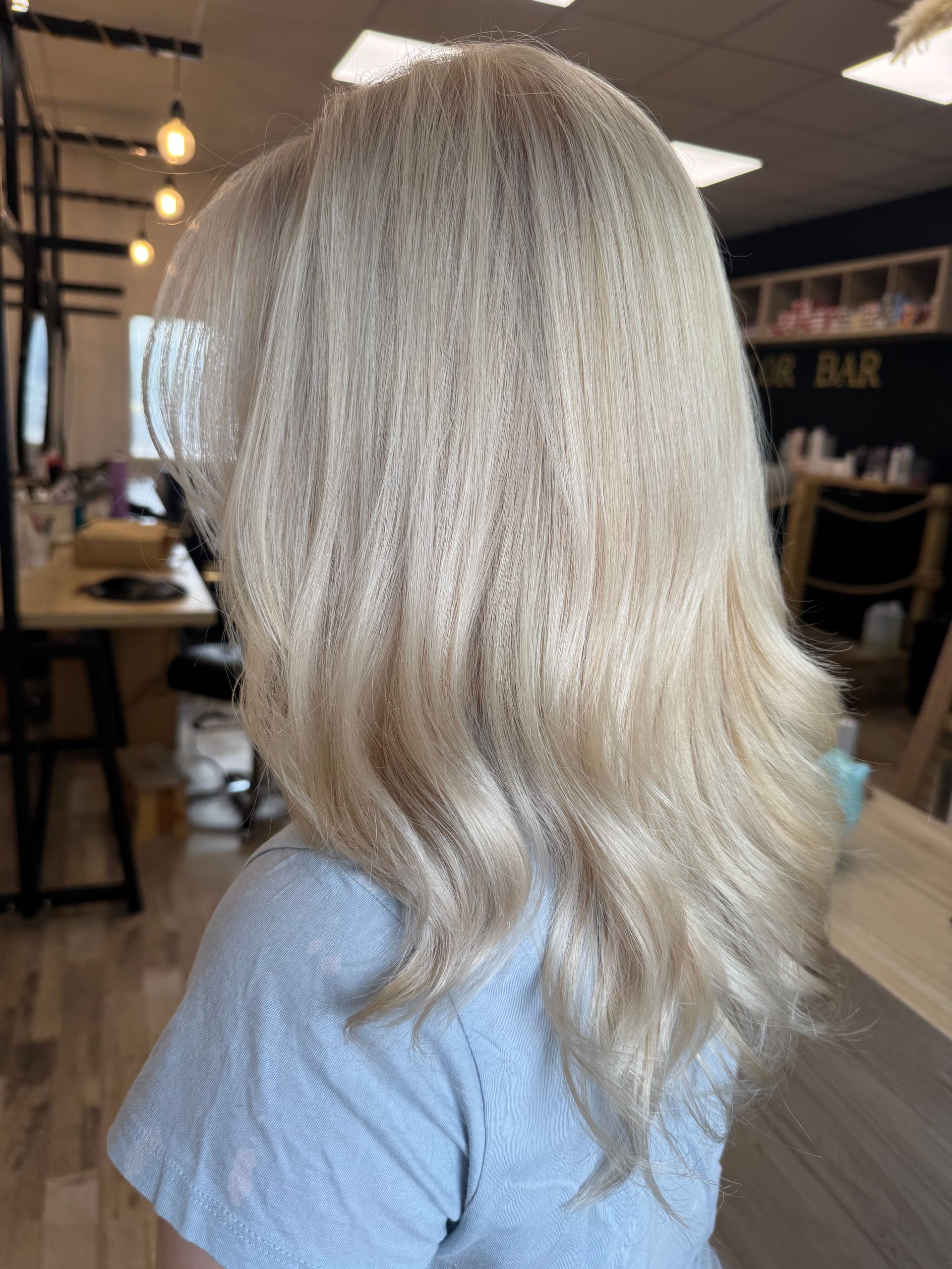 Cheveux blonds ondulés vus de dos dans un salon, avec un haut bleu clair et des lumières chaleureuses en arrière-plan
