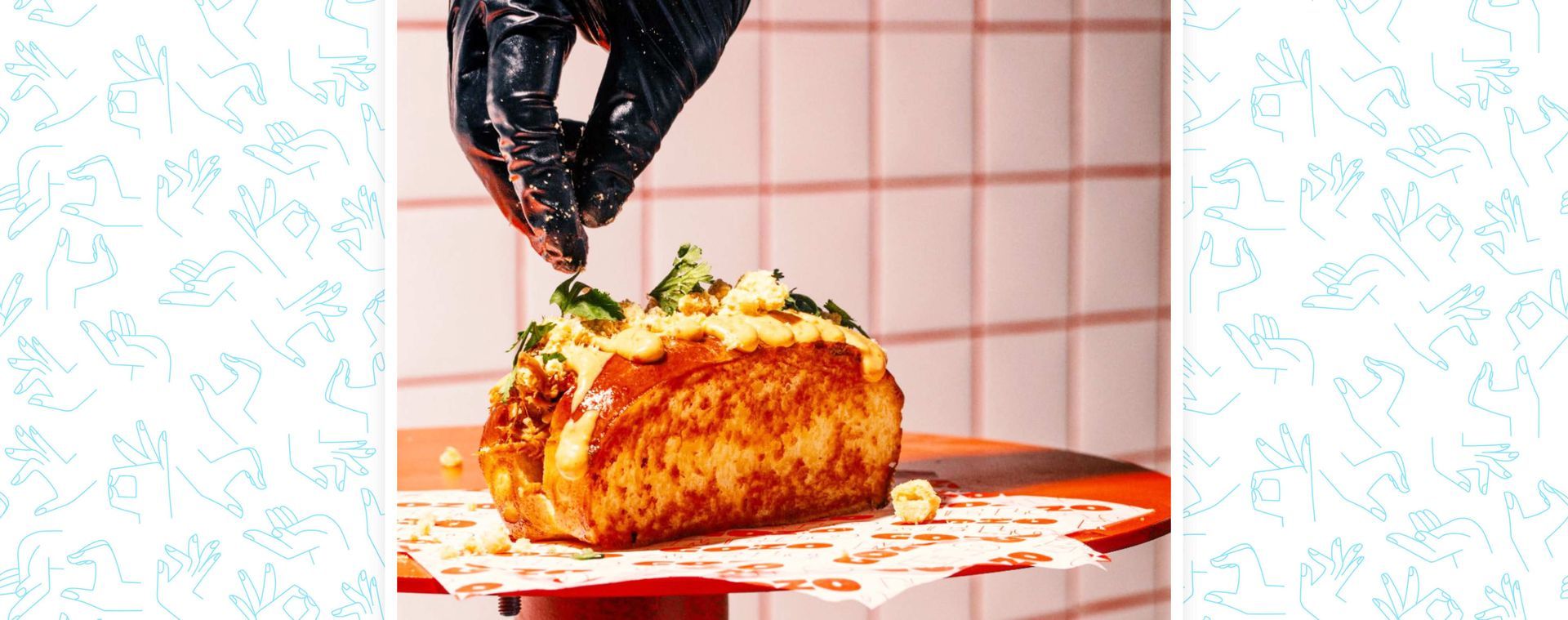 Una mano enguantada espolvorea guarnición sobre un sándwich tostado servido en una bandeja forrada con papel, 