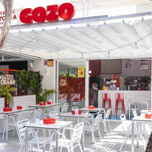 Exterior del restaurante con mesas al aire libre: mesas blancas, sillas rojas, cartel