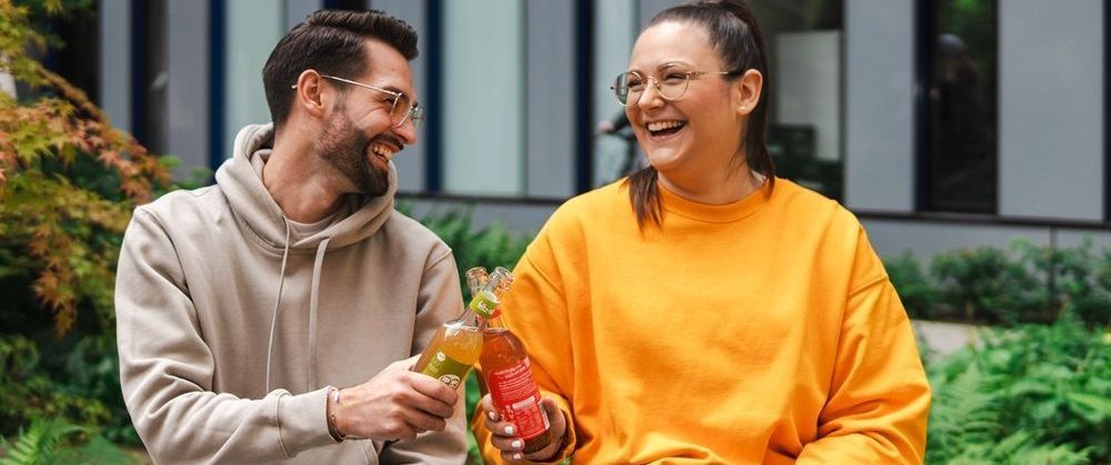 Zwei Personen stoßen mit Getränken an. Der Mann trägt einen Kapuzenpullover, die Frau einen orangefarbenen Pullover.