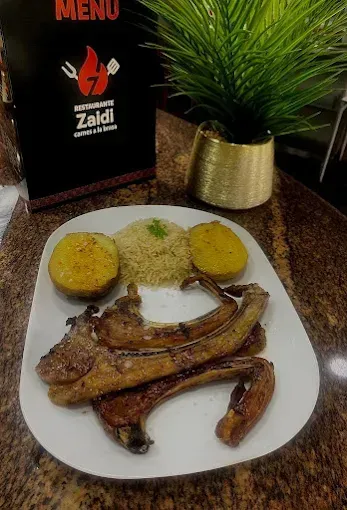 Un plato blanco con chuletas de cordero a la parrilla, arroz y dos mitades de patata asads.