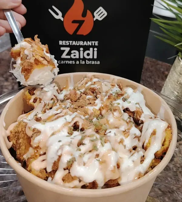 Una mano sosteniendo una cuchara llena de un plato de comida frita y con salsa del Restaurante Zaidi.