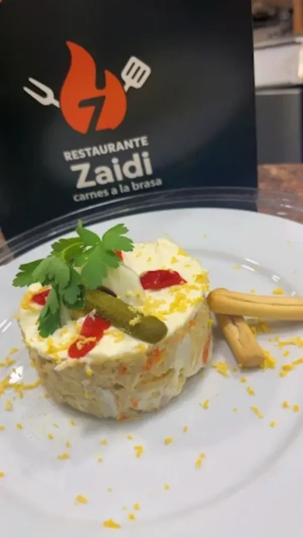 Un plato con una porción de ensalada rusa, aderezada con perejil y un pepinillo, con el logotipo del Restaurante Zaidi.