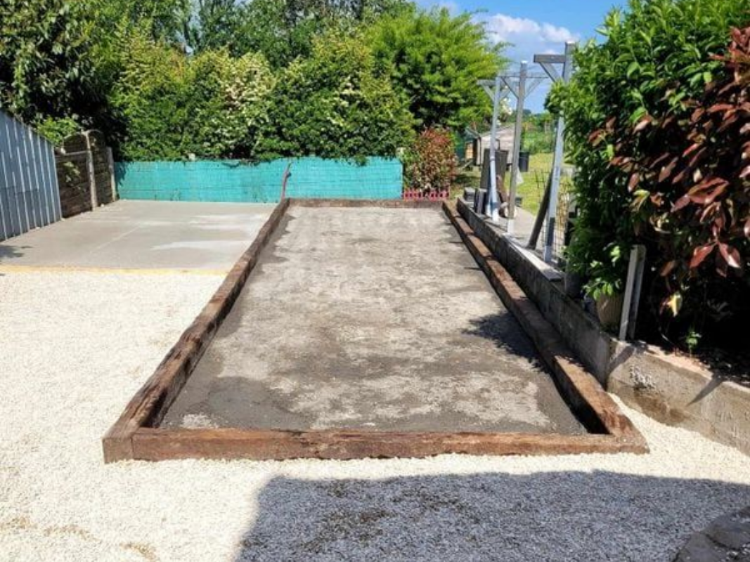 Un terrain de pétanque rectangulaire aux bordures en bois, entouré de gravier et de verdure.