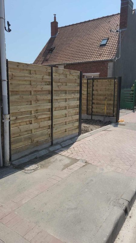 Clôture en bois à cadre noir, trottoir gris, bâtiment en briques en arrière-plan par une journée ensoleillée.