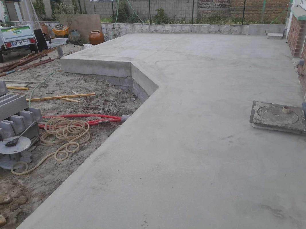 Une terrasse en béton est en construction à l'extérieur ; on aperçoit du sable, des matériaux et un mur de briques.