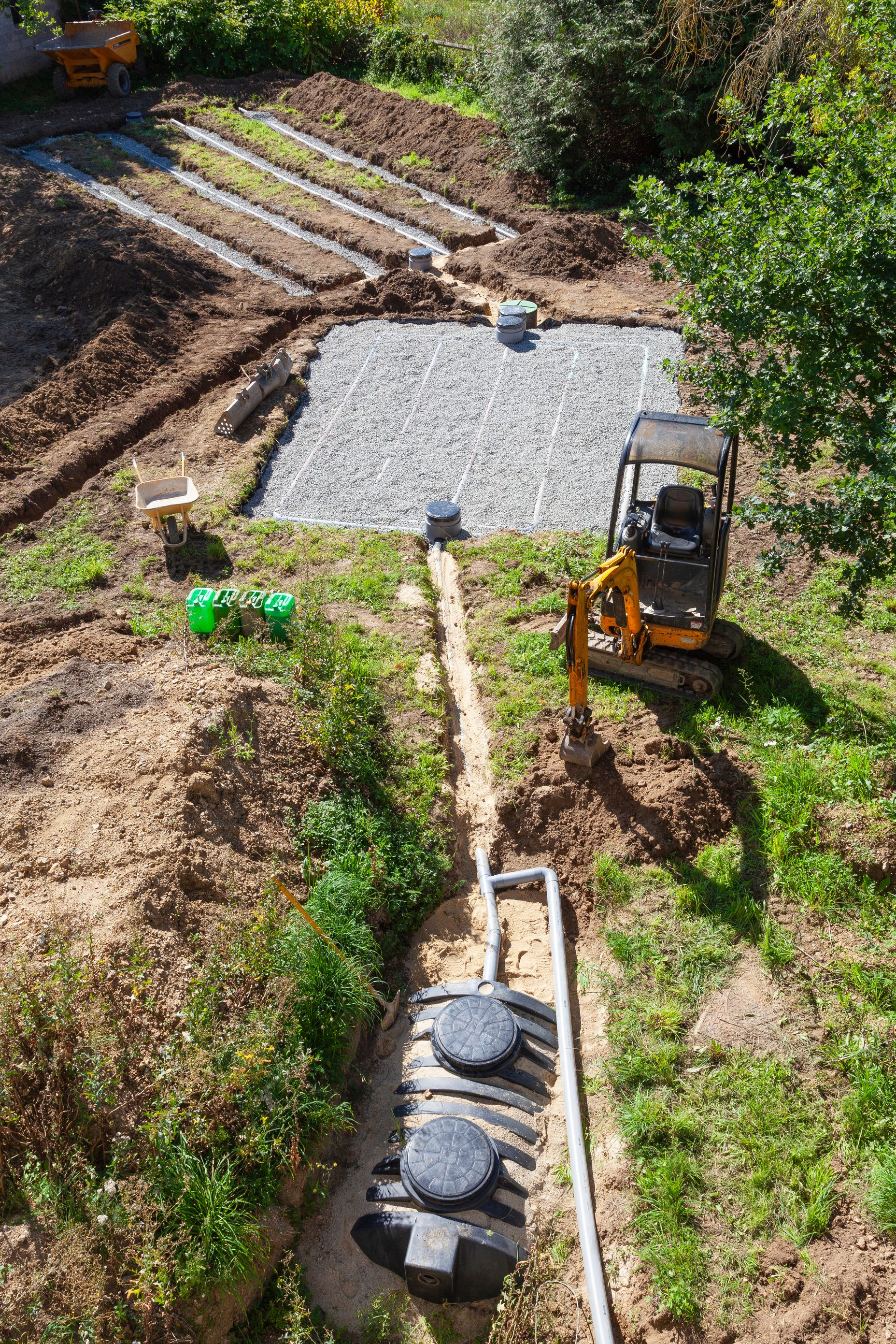 Chantier de construction avec installation de fosse septique : excavatrice, lit de gravier, tuyaux et réservoirs dans une zone herbeuse.