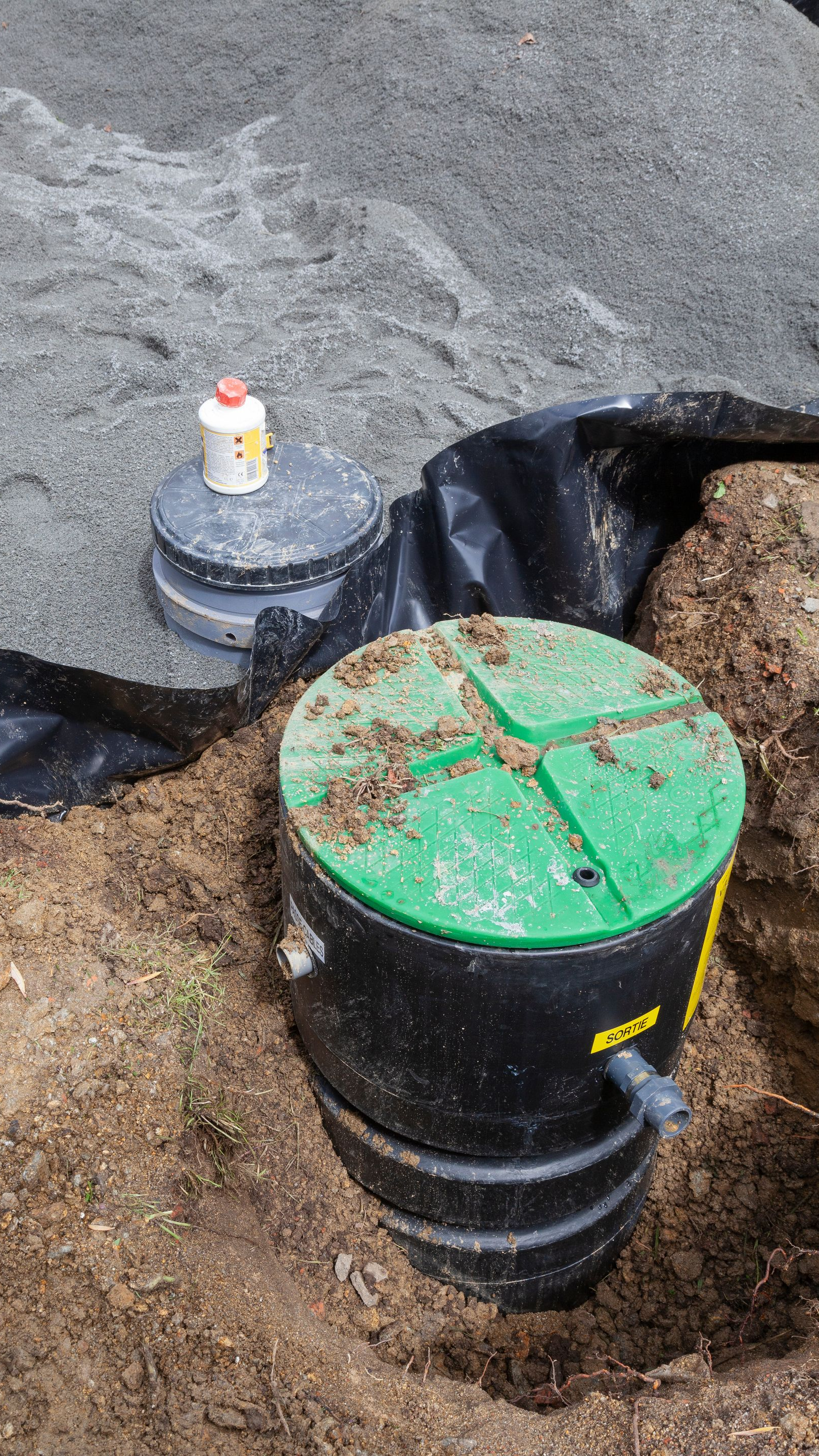 Deux fosses septiques noires à couvercles verts sont installées dans un trou de terre. Un tas de gravier se trouve à l'arrière-plan.