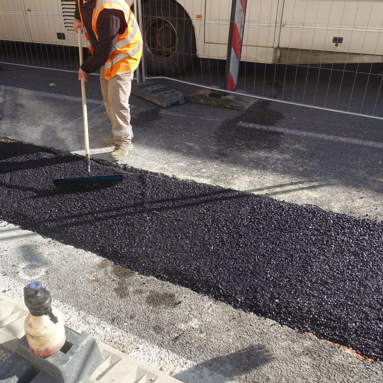 Un ouvrier routier en gilet orange utilise un râteau sur un revêtement d'asphalte frais, près d'un bus.