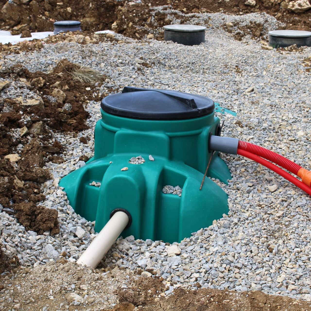 Fosse septique verte installée dans du gravier, avec couvercle noir, tuyaux et terre environnante.