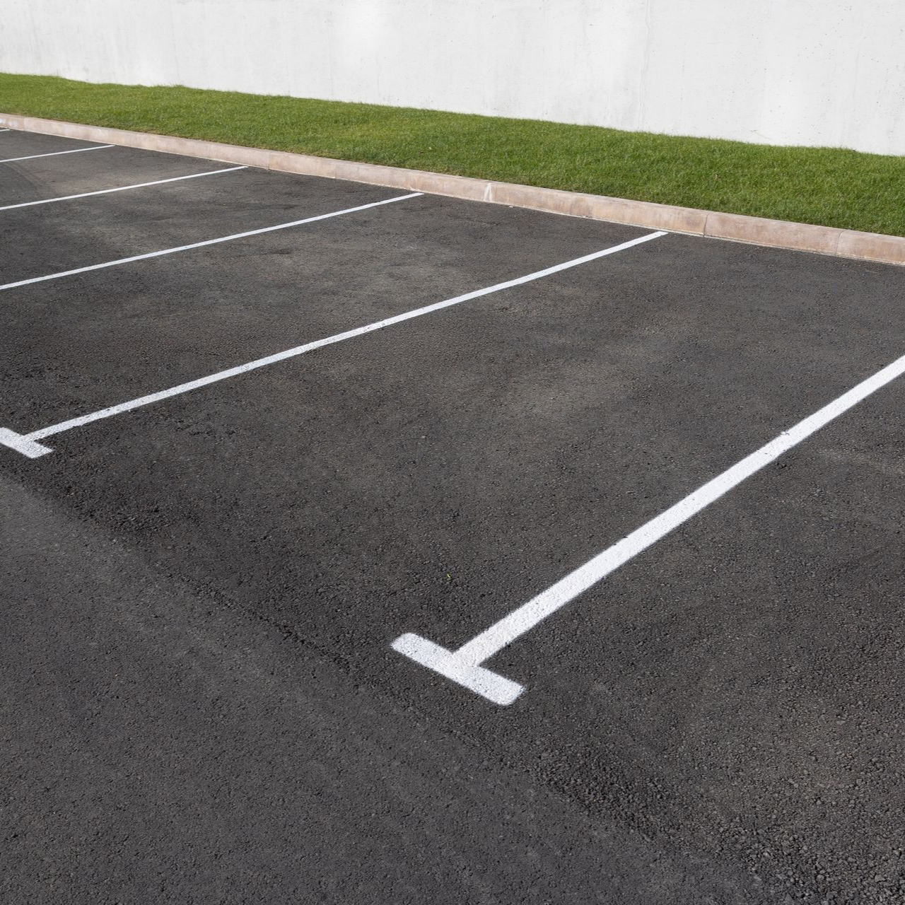 Places de parking délimitées par de la peinture blanche sur l'asphalte noir ; bordure de gazon le long d'un mur blanc.