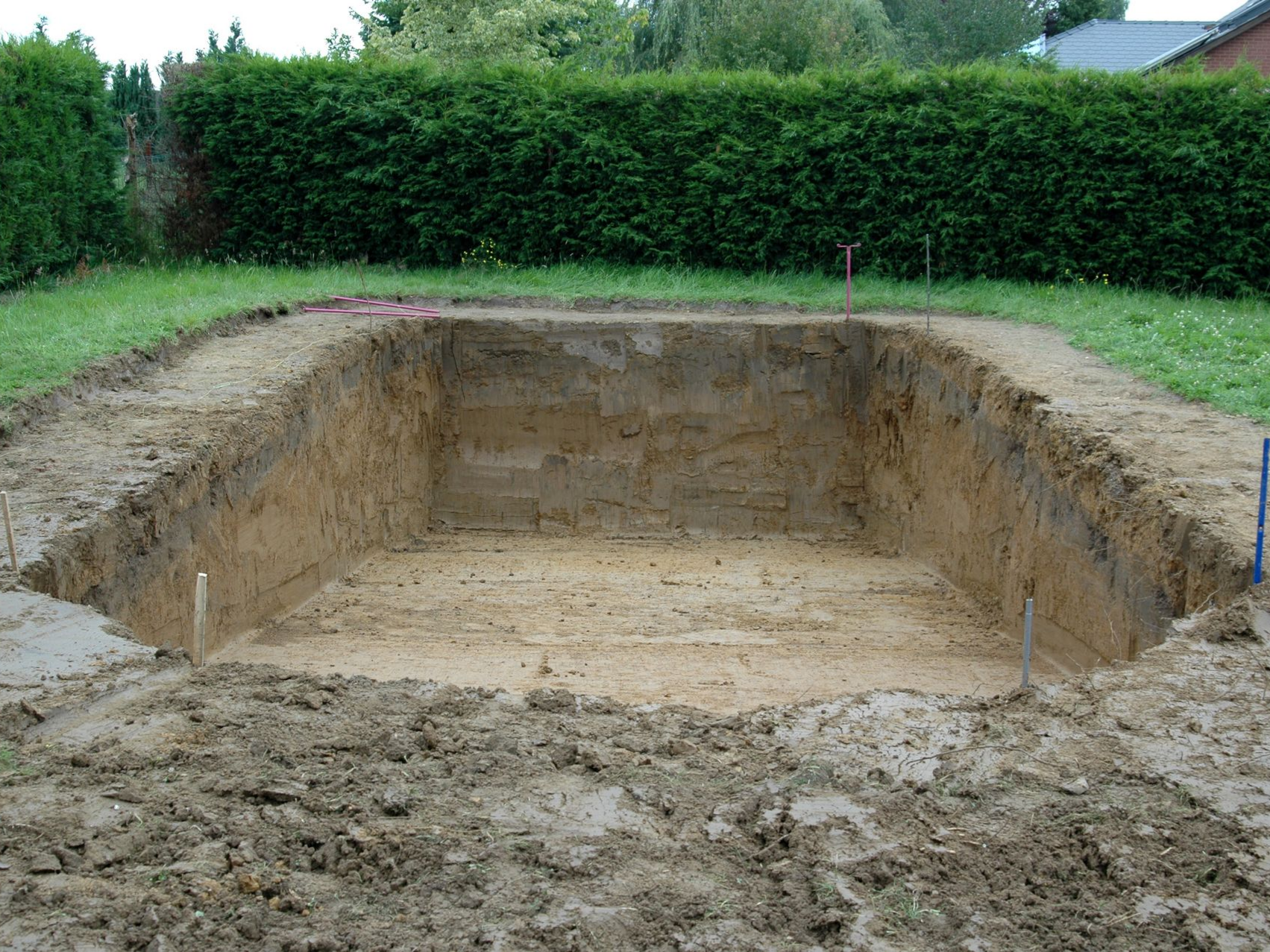 Trou rectangulaire creusé dans le sol, probablement pour une piscine, entouré de boue et d'herbe.