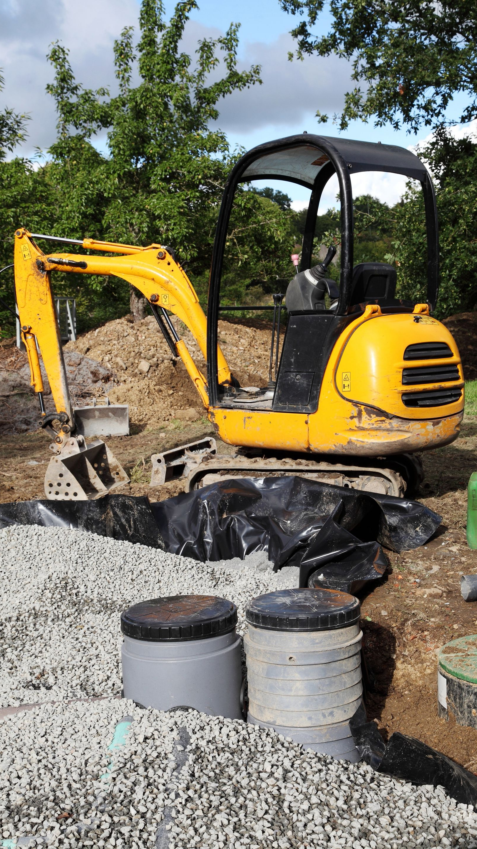 Une pelleteuse jaune travaille sur un chantier de construction avec du gravier, des tuyaux et un marquage noir.