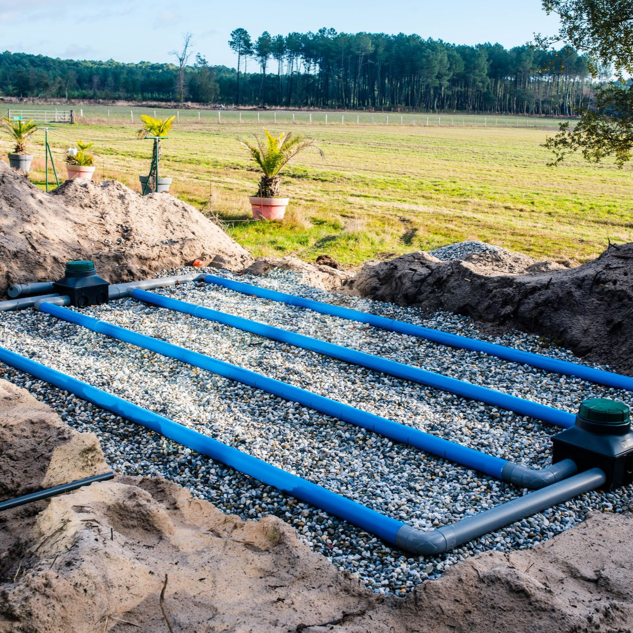 Champ d'épandage septique souterrain avec tuyaux bleus, gravier et champ vert en arrière-plan.