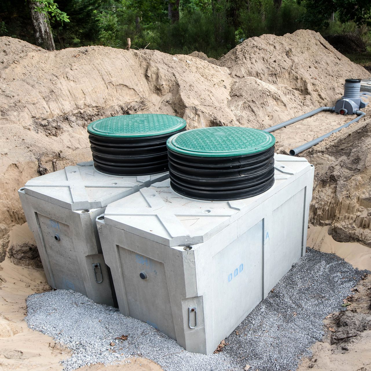 Deux fosses septiques en béton avec couvercles verts, partiellement enterrées dans une zone sablonneuse, gravier à proximité.