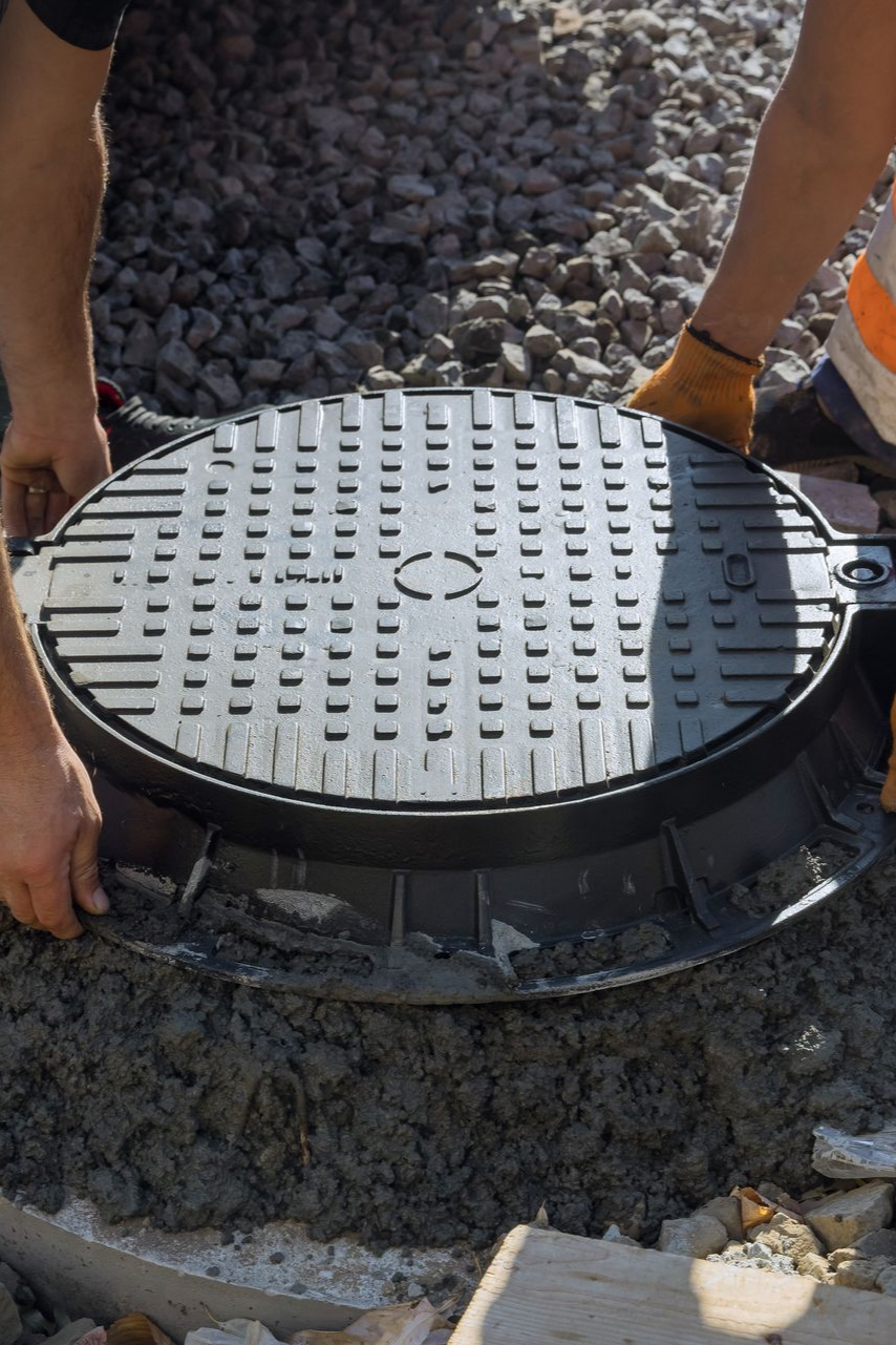 Des ouvriers installent une plaque d'égout noire sur une surface de gravier.